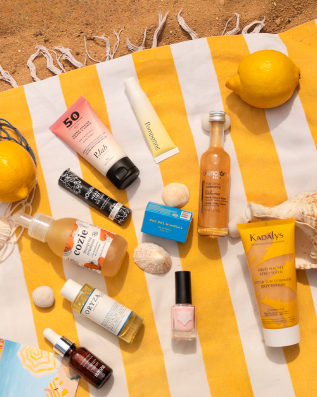 Admirez les produits naturels de la box French Riviera disposés sur une serviette rayée à la plage, incluant soins solaires SPF 50, huiles sublimes, nettoyants doux et maquillage pétillant pour l’été.